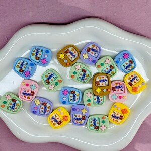 May include: A collection of colorful, miniature game console-shaped embellishments. Each charm features a screen with a bear graphic and the word "Happy". The charms are in various colors, including blue, pink, yellow, and green, and are arranged on a white, wavy-edged plate.
