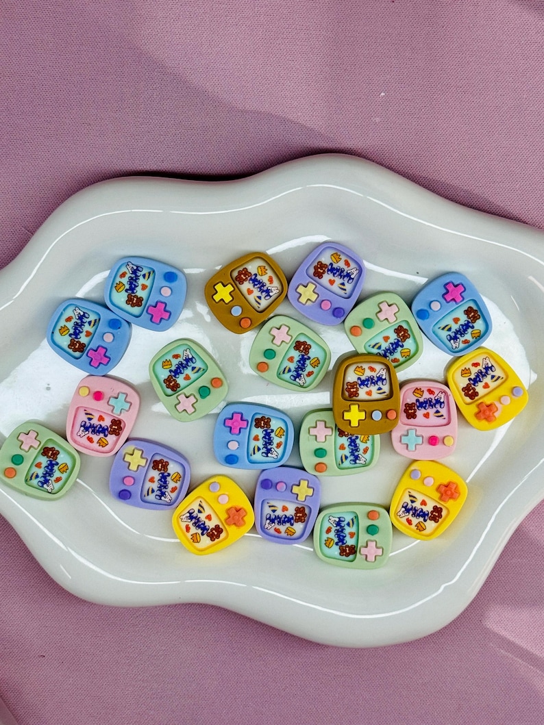May include: A collection of colorful, miniature game console-shaped embellishments. Each charm features a screen with a bear graphic and the word "Happy". The charms are in various colors, including blue, pink, yellow, and green, and are arranged on a white, wavy-edged plate.