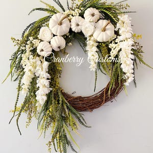 May include: A decorative wreath featuring a brown twig base adorned with white pumpkins, white floral clusters, and various green foliage. The text Cranberry Customs is written in white script across the centre of the arrangement.