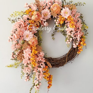 May include: A circular wreath featuring a brown twig base adorned with an arrangement of artificial flowers in shades of soft pink, peach, and golden yellow, accented with green foliage and the text Cranberry Customs written in white script.