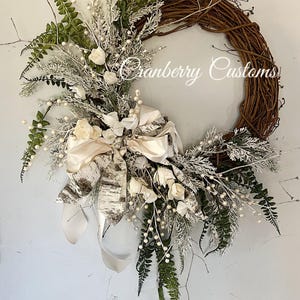 Peut inclure: Une couronne de vigne artisanale ornée de roses blanches, de verdure artificielle et d'un nœud crème avec des accents d'écorce de bouleau. La couronne présente de délicates baies blanches et des brins.