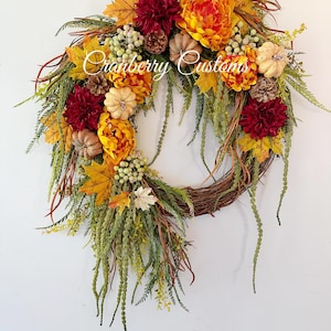Op de afbeelding: Een decoratieve krans met een basis van gevlochten takken, versierd met donkerrode dahlia's, feloranje pioenen, kleine beige pompoenen, dennenappels, groene bessen en diverse herfstkleurige esdoornbladeren. De tekst Cranberry Customs is zichtbaar.
