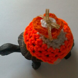 May include: A small turtle wearing a crocheted orange and white sweater with a gold ribbon bow on top.