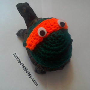 May include: A turtle wearing a handmade green and orange crocheted costume. The costume includes an orange headband with white and black eye accents. The text "loolapets@etsy.com" is visible.