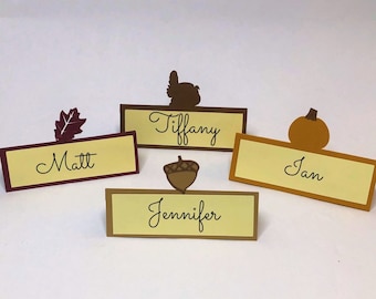 May include: Four place cards with fall-themed designs. The place cards are yellow with brown borders and feature the names Tiffany, Matt, Jan, and Jennifer.