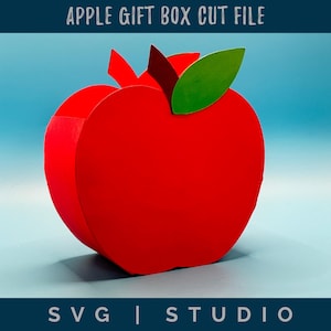 May include: A red apple-shaped gift box cut file. The box has a green leaf and a stem. The text "APPLE GIFT BOX CUT FILE" is at the top. The text "SVG | STUDIO" is at the bottom. The background is a gradient of blue.