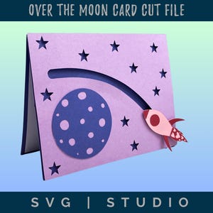 Op de afbeelding: Een lavendelkleurige kaartuitsnede met een ruimtethema. De kaart heeft een donkerblauwe maan met witte kraters, een raket en steruitsparingen. De tekst "OVER THE MOON CARD CUT FILE" en "SVG | STUDIO" staan bovenaan en onderaan.
