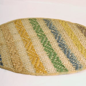 May include: Oval-shaped woven mat with diagonal stripes in shades of beige, yellow, green, blue, and brown. The mat is made of natural fibers and has a textured surface. The edges are finished with a woven border.