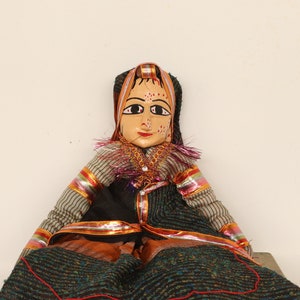 May include: A handcrafted wooden doll with a painted face, wearing a black and teal patterned dress. The doll has a painted face with red lips and a decorative headpiece. The doll's arms are striped with red and gold accents. The doll is sitting.