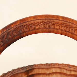 May include: Close-up of a wooden basket handle with intricate carved floral and leaf patterns. The handle is a rich, warm brown color, and the carving details are clearly visible. The basket's design suggests a rustic or vintage aesthetic.