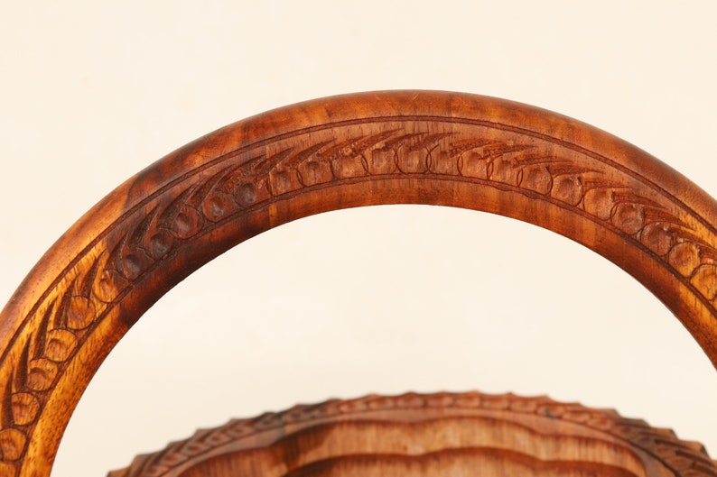 May include: Close-up of a wooden basket handle with intricate carved floral and leaf patterns. The handle is a rich, warm brown color, and the carving details are clearly visible. The basket's design suggests a rustic or vintage aesthetic.