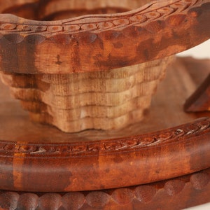 May include: Close-up of a handcrafted wooden fruit bowl. The bowl features intricate carved details and a tiered design. The wood has a rich, dark brown color, with lighter tones in the center. The bowl is likely used for decorative purposes.