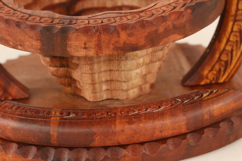 May include: Close-up of a handcrafted wooden fruit bowl. The bowl features intricate carved details and a tiered design. The wood has a rich, dark brown color, with lighter tones in the center. The bowl is likely used for decorative purposes.