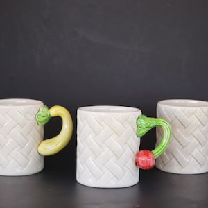 Könnte beinhalten: Drei weiße Keramikbecher mit einer gewebten Textur. Jeder Becher hat einen einzigartigen Griff in Form eines Gemüses: eine gelbe Zucchini, eine rote Tomate und eine orangefarbene Karotte. Die Griffe sind mit grünen Blättern versehen.