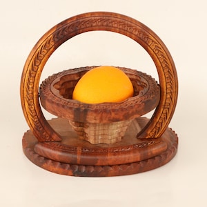 May include: A handcrafted wooden fruit basket with an orange inside. The basket is made of dark brown wood and features intricate carved designs. The basket has a circular base and a handle that arches over the top.