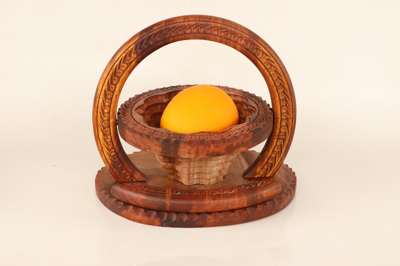 May include: A handcrafted wooden fruit basket with an orange inside. The basket is made of dark brown wood and features intricate carved designs. The basket has a circular base and a handle that arches over the top.