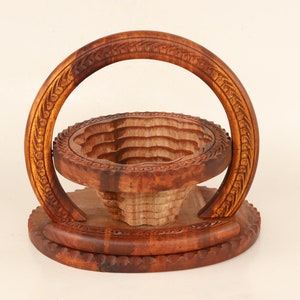 May include: A handcrafted wooden fruit basket with a carved design. The basket features a round base, a tiered bowl, and a curved handle, all made of warm brown wood. The handle and rim have intricate floral carvings.