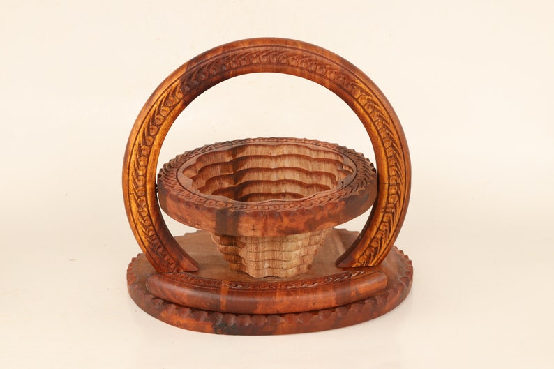 May include: A handcrafted wooden fruit basket with a carved design. The basket features a round base, a tiered bowl, and a curved handle, all made of warm brown wood. The handle and rim have intricate floral carvings.