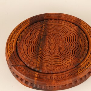 May include: A round, wooden fruit basket with a carved floral design in the center. The basket is made of dark brown wood and has a collapsible design. The rim of the basket has a decorative carved pattern.
