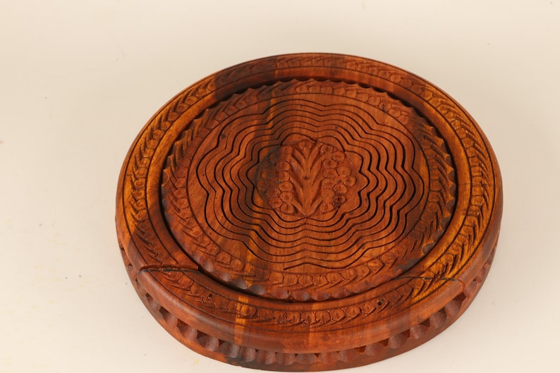 May include: A round, wooden fruit basket with a carved floral design in the center. The basket is made of dark brown wood and has a collapsible design. The rim of the basket has a decorative carved pattern.