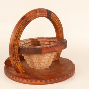 May include: A handcrafted wooden basket with a unique design. The basket features a round base, a tiered bowl, and a curved handle. The wood has a warm brown color, with carved details on the handle and rim. The basket is likely used for decorative or storage purposes.