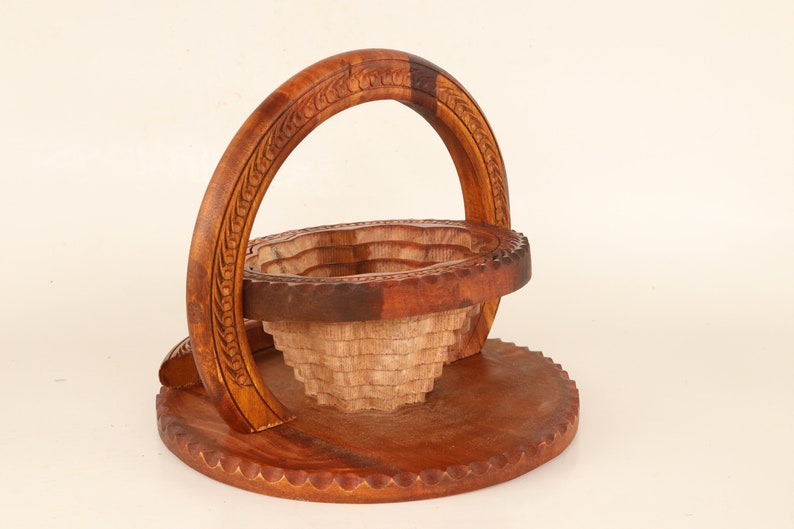 May include: A handcrafted wooden basket with a unique design. The basket features a round base, a tiered bowl, and a curved handle. The wood has a warm brown color, with carved details on the handle and rim. The basket is likely used for decorative or storage purposes.