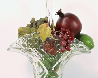 May include: A clear glass basket filled with artificial fruit, including green and red grapes, a pomegranate, and a lime. The basket has a decorative, textured pattern and a handle. The arrangement is set against a white background.