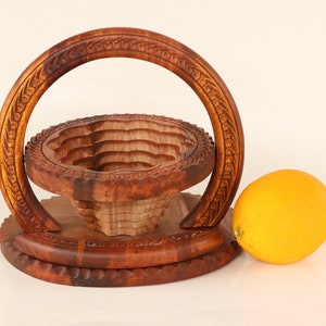 May include: A handcrafted wooden fruit basket with a carved design, featuring a circular handle and a tiered, expandable bowl. An orange sits beside the basket. The basket is made of a warm, reddish-brown wood.
