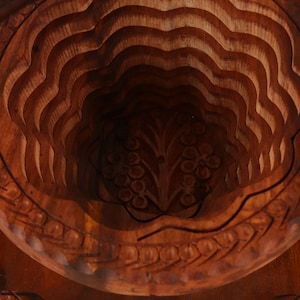 May include: A detailed, close-up view of a carved wooden fruit basket. The basket is made of dark brown wood and features a layered, wave-like design. Intricate floral patterns are carved into the base and rim, adding to its decorative appeal.