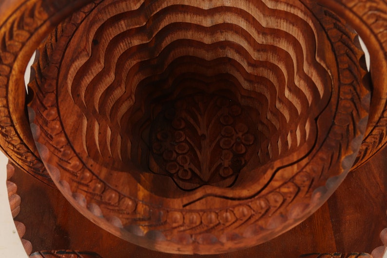 May include: A detailed, close-up view of a carved wooden fruit basket. The basket is made of dark brown wood and features a layered, wave-like design. Intricate floral patterns are carved into the base and rim, adding to its decorative appeal.