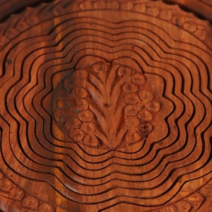 May include: Close-up of a wooden fruit basket with a carved floral design in the center. The basket is made of layered, wavy wooden slats, and the wood has a warm, reddish-brown color.