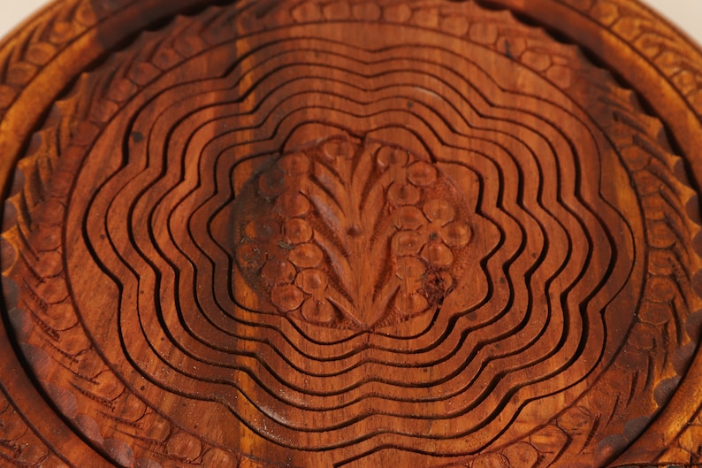 May include: Close-up of a wooden fruit basket with a carved floral design in the center. The basket is made of layered, wavy wooden slats, and the wood has a warm, reddish-brown color.