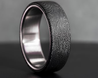 May include: A dark gray metal ring featuring a complex, etched fingerprint-like pattern on the outer surface. The band has a smooth, polished silver-toned interior finish, creating a distinct contrast between the textured exterior and the metallic inner band.