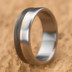 Modern men's engagement ring in titanium and carbon fiber with brass accent, standing on tan stone surface