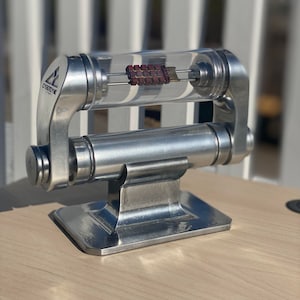 Puede incluir: Un soporte de metal plateado con un tubo transparente que contiene un pequeño objeto rojo y dorado. El soporte tiene la palabra "CYBERZONE" impresa.