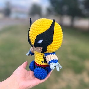 May include: A crocheted plush toy of Wolverine from the X-Men. The toy is yellow with a black mask and blue pants. The toy is being held in a person's hand.