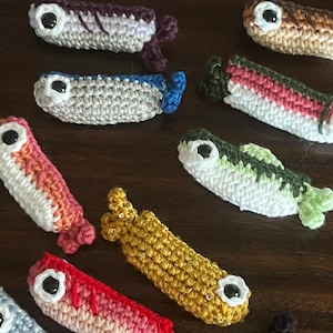 May include: A collection of small, handmade crochet fish ornaments in various colors including gold, red, green, blue, and brown. Each fish features a prominent black plastic eye, textured yarn stitching, and a small tail fin at the end.