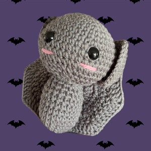 May include: A grey crocheted bat plush toy with black eyes and pink cheeks. The bat is sitting on a purple background with a pattern of black bats.