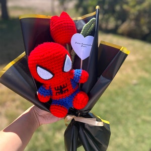 May include: A crocheted Spiderman figure with a red and blue suit, holding a red crocheted flower. The bouquet is wrapped in black paper with a gold ribbon and a white heart-shaped tag that says "Feliz Cumplea&ntilde;os Peter".
