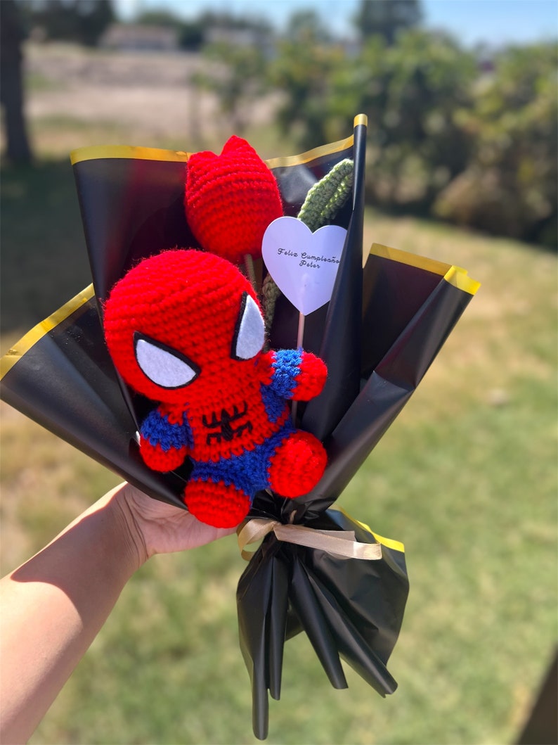 May include: A crocheted Spiderman figure with a red and blue suit, holding a red crocheted flower. The bouquet is wrapped in black paper with a gold ribbon and a white heart-shaped tag that says "Feliz Cumplea&ntilde;os Peter".