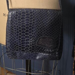 May include: A dark blue crossbody bag featuring a textured, reptilian-inspired pattern across the flap and body. The bag includes a long, matching shoulder strap and a small, rectangular embossed patch on the lower right corner of the front flap.