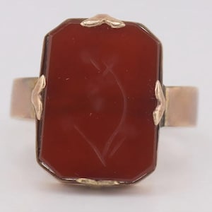 May include: A gold ring featuring a large, rectangular, deep red carnelian gemstone set in a metal frame. The stone has a subtle, etched floral design on its surface, held securely by four small, decorative metal prongs at the corners.