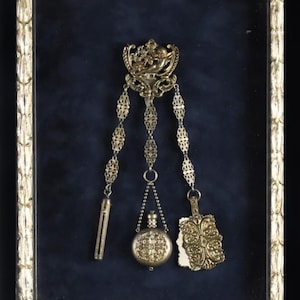 May include: A gold-toned chatelaine brooch featuring an ornate top piece with a cherub motif, connected by three delicate filigree chains to a cylindrical needle case, a small round perfume bottle, and a rectangular embossed decorative tablet.