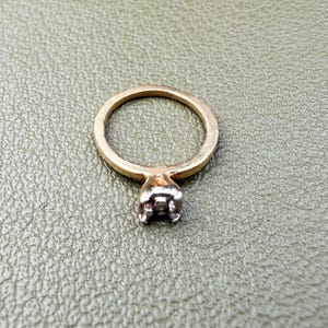 May include: A gold ring featuring a silver-toned metal setting designed to hold a single stone. The band is a simple, smooth gold circle that connects to a four-prong mounting at the top, which is currently empty of any gemstone or setting.