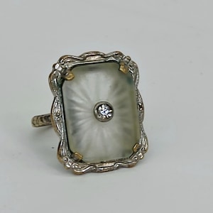 May include: A rectangular ring featuring a frosted glass center with a sunburst pattern and a single clear stone set in the middle. The glass is held by a silver-toned metal frame with ornate, scalloped edges and a textured band design.