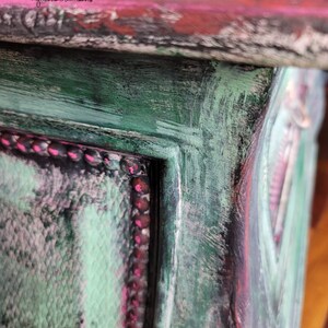 May include: Close-up of a wooden box with a distressed paint finish. The box is painted in shades of green, pink, and black. The edges of the box are highlighted with a pink trim.