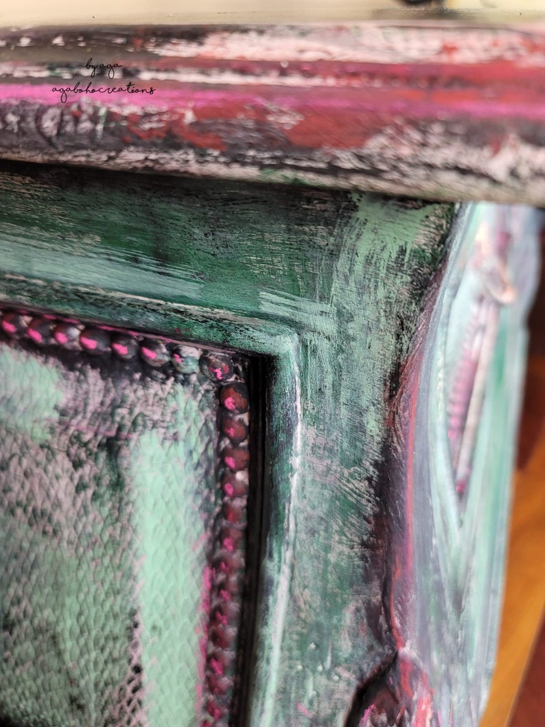 May include: Close-up of a wooden box with a distressed paint finish. The box is painted in shades of green, pink, and black. The edges of the box are highlighted with a pink trim.