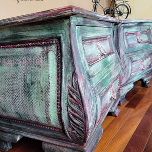 May include: Two antique green painted bedside tables with ornate carvings and distressed details. Each table has three drawers with metal handles. The tables are sitting on a wooden floor.