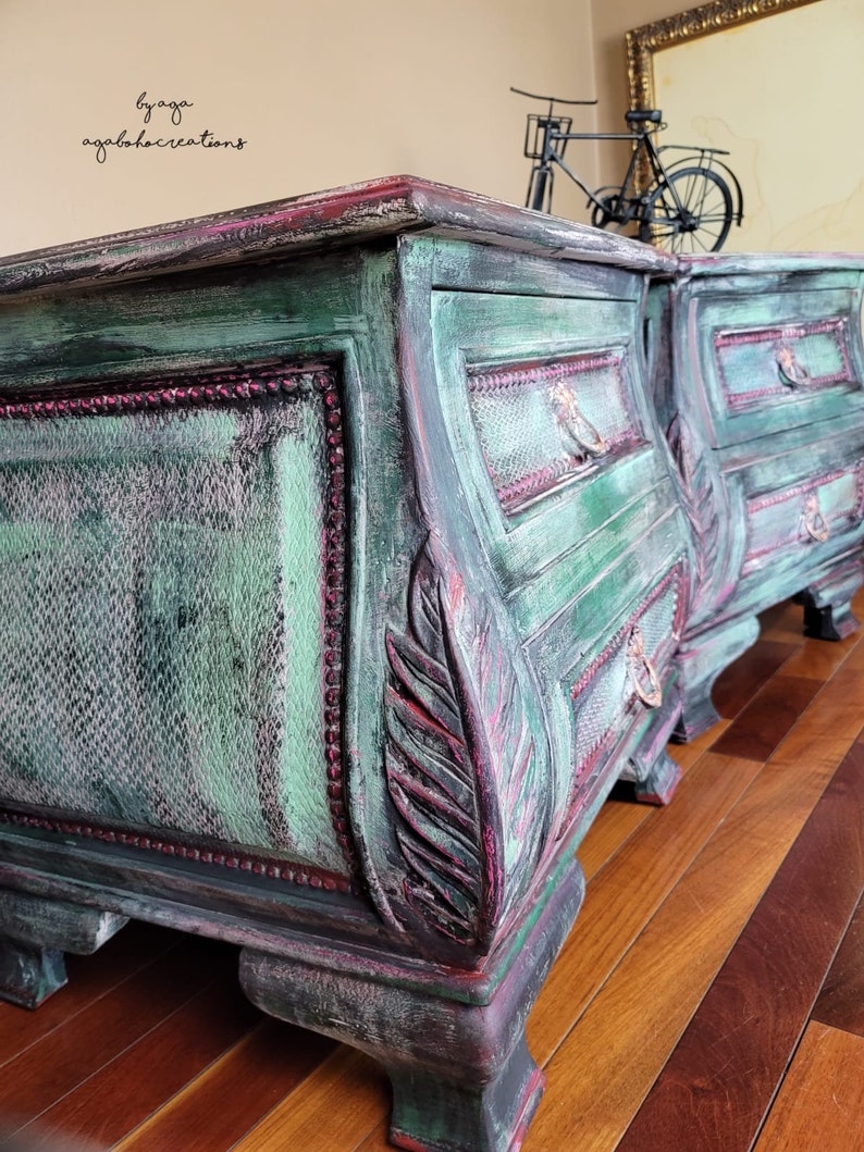 May include: Two antique green painted bedside tables with ornate carvings and distressed details. Each table has three drawers with metal handles. The tables are sitting on a wooden floor.