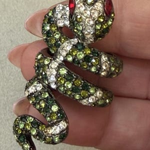 May include: A coiled snake ring featuring a dark metal base encrusted with numerous small, sparkling rhinestones in shades of lime green, forest green, and clear white. The snake has two prominent, round red stones serving as its eyes.